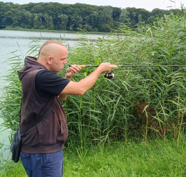 Mann angelt am Ufer eines Gewässers, umgeben von hohem Schilf.